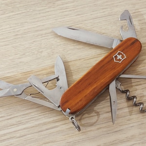 Peut inclure: Un couteau de poche multifonction avec des plaquettes en bois poli et un emblème argenté en forme de croix et de bouclier. L'outil en acier inoxydable présente une lame principale ouverte, des ciseaux, un décapsuleur, un tire-bouchon et un crochet.