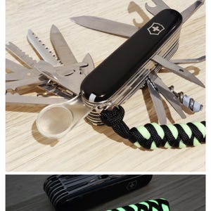 Può includere: Un coltellino svizzero multifunzione nero con numerosi utensili in acciaio inossidabile estratti, inclusa una lente d'ingrandimento. Un cordino in paracord intrecciato nero e verde neon è fissato all'anello metallico alla base del manico.
