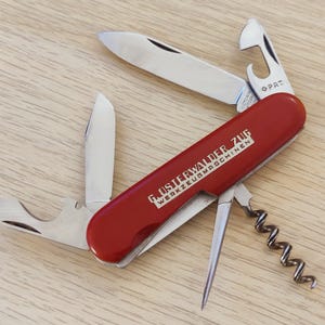 Puede incluir: Una navaja multiusos roja con varias hojas y herramientas de acero inoxidable desplegadas. El mango presenta el texto impreso en blanco G. OSTERWALDER ZUG WERKZEUGMASCHINEN. Las herramientas incluyen una hoja principal, una hoja pequeña, un abridor de botellas y un sacacorchos.