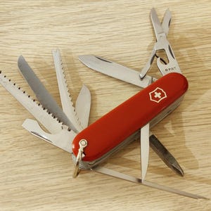 Può includere: Un coltellino svizzero multifunzione rosso con un emblema a croce bianca sul manico. Diversi utensili in acciaio inossidabile sono aperti, tra cui una sega, una lama, forbici, un apribottiglie e un cacciavite, con un anello in metallo.