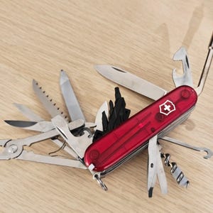 Könnte beinhalten: Ein rotes Multifunktions-Taschenmesser mit transparentem Griff und weißem Kreuz-Emblem. Das Werkzeug ist vollständig ausgeklappt und zeigt verschiedene Edelstahl-Elemente wie Zange, Säge, Klinge, Korkenzieher und einen Satz Schraubendreher-Bits.