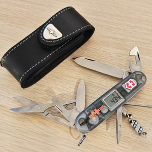 Könnte beinhalten: Ein Multifunktions-Taschenmesser mit einem durchscheinenden grauen Griff, einer Digitalanzeige mit der Zahl 434 und dem Rega-Logo. Es verfügt über mehrere Edelstahlklingen, eine Schere, einen Korkenzieher und einen Flaschenöffner sowie ein schwarzes Lederetui.