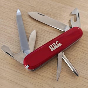 Op de afbeelding: Een rood multifunctioneel zakmes met de witte tekst BBC BROWN BOVERI op het handvat. Het gereedschap wordt getoond met verschillende roestvrijstalen onderdelen uitgeklapt, waaronder een hoofdmes, een nagelvijl, een flesopener en een schroevendraaier.