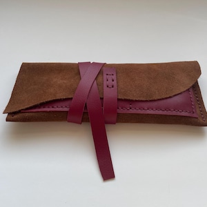 May include: A handmade leather case in shades of brown and burgundy. The case is rectangular with a flap closure and a burgundy leather strap that wraps around it. The stitching is visible, and the leather has a textured appearance.