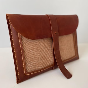 Pode incluir: Uma clutch de couro castanho com fecho de aba e um painel de tecido de espinha de peixe tecido. A clutch apresenta uma alça de couro com fivela e é costurada com linha contrastante.