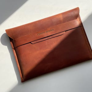 Puede incluir: Una funda de cuero marrón con cierre de solapa. La funda tiene un borde cosido y el texto "CEREN AKMAN" está grabado en la parte delantera. El cuero tiene un grano natural y un color marrón rojizo.