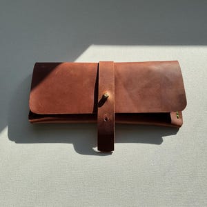 May include: A brown leather clutch with a strap closure. The clutch is rectangular with rounded corners and a flap that folds over. The strap is secured with a brass-colored stud. The leather has a natural, textured appearance.
