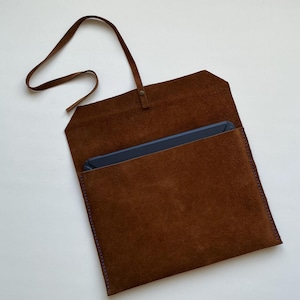 Puede incluir: Una funda de tableta de gamuza marrón con una correa a juego y un cierre de botón. El interior está forrado con un material más oscuro y los bordes están cosidos con hilo morado. La funda está abierta, revelando una tableta en el interior.