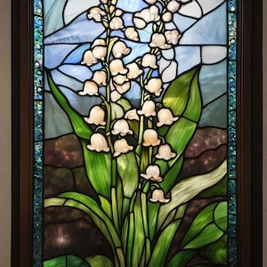 Puede incluir: Una ventana de vitrales que representa un ramo de flores blancas de lirio de los valles con hojas verdes sobre un fondo de cielo azul y blanco.