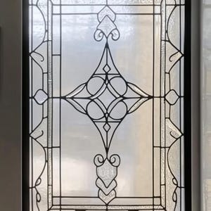 Może przedstawiać: Witraż z geometrycznym wzorem w kształcie rombu z misternymi spiralami i zakrzywieniami. Okno jest oprawione w czerni.