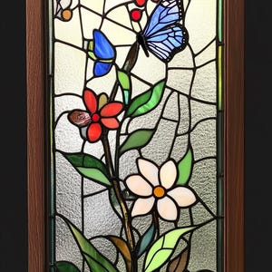 May include: A stained glass window panel featuring a floral design with butterflies. The panel depicts a white flower with a yellow center, a red flower with a yellow center, and two blue butterflies. The background is a light blue with a white border.