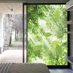 Puede incluir: Una ventana grande con un diseño vibrante de hojas verdes, creando una escena natural al aire libre. La imagen muestra una vista de un bosque a través de la ventana, con la luz del sol que atraviesa las hojas. La ventana está enmarcada en negro, y el interior tiene una cama y estanterías.