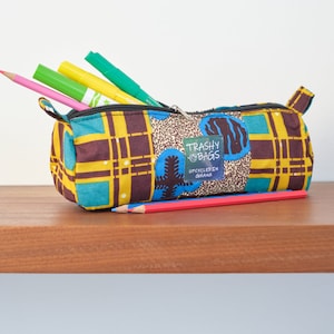 May include: A yellow, brown, blue, and green patterned pencil case with a zipper closure. The case is filled with colored pencils and markers. The case has a label that reads "Trashy Bags" and "Upcycled in Ghana".
