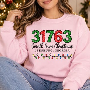 May include: A light pink crewneck sweatshirt featuring the number 31763 in green and red with snowflake patterns, the text Small Town Christmas and LEESBURG, GEORGIA, and a graphic of a string of colorful Christmas lights below.