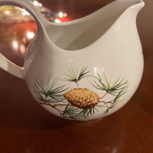 May include: White ceramic creamer with a pine cone and pine needles design. The creamer has a rounded body and a handle.