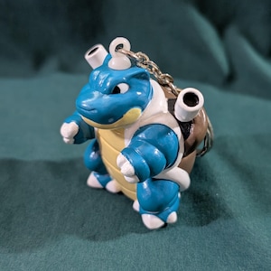 May include: A blue, white, and tan Blastoise figurine with a silver keychain. The figurine has a detailed design, featuring the character's signature features. The figure is approximately 3 inches tall.