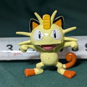 May include: A yellow Meowth figurine with orange accents on its feet and tail. The figure has a smiling expression and is posed with its arms outstretched. A measuring tape is in the background.