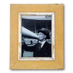 May include: A framed black and white photograph of a man speaking into a megaphone. The frame is rectangular with a distressed yellow finish and a white inner border. The image captures a vintage scene.