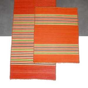 Op de afbeelding: Twee geweven placemats in oranje en wit met strepen. De grotere placemat heeft een breder strepenpatroon en de kleinere placemat heeft een smaller strepenpatroon.