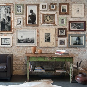 Könnte beinhalten: Eine Galeriewand mit einer Sammlung gerahmter Schwarz-Weiß-Fotografien in verschiedenen Größen. Die Rahmen sind in verschiedenen Stilen und Farben, darunter Weiß, Braun und Grün, mit einem Used-Look. Vor der Wand steht ein grüner Tisch mit Büchern und Dekoration.