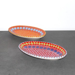 May include: Two oval ceramic serving dishes with colorful geometric patterns. The dish on the left has a red, orange, and yellow pattern. The dish on the right has a red, blue, and yellow pattern.