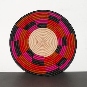 Könnte beinhalten: Ein runder gewebter Wandbehang mit einem geometrischen Muster in Schwarz, Rot, Orange und Pink. Die Mitte des Wandbehangs ist ein natürlicher gewebter Kreis.