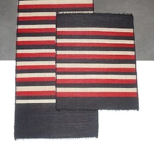 May include: Three rectangular rugs with a striped pattern. The rugs feature alternating bands of black, red, and off-white. The rugs are made of a woven material and have a textured appearance. The rugs are arranged in a layered composition.