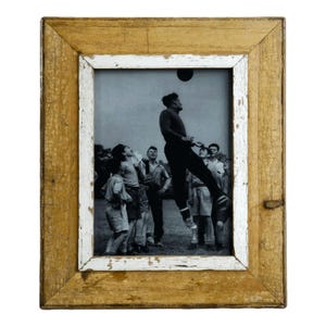May include: A framed black and white photograph of a group of people playing a sport. The frame is made of distressed wood with a white inner border. The photograph shows a person jumping to hit a ball.