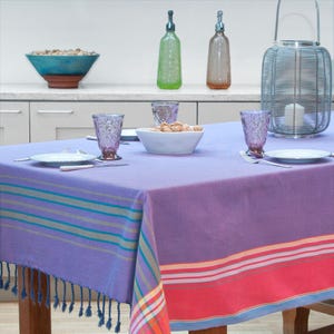 Op de afbeelding: Een lavendelkleurig tafelkleed met kleurrijke gestreepte randen en franjes. De tafel is gedekt met borden, glazen en bestek. Decoratieve glazen flessen en een metalen lantaarn staan op de achtergrond. Een kom met noten staat op tafel.