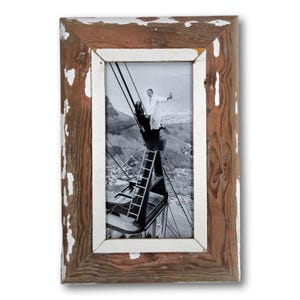 May include: Rectangular wooden picture frame with a distressed, rustic look. The frame has a brown finish with white paint chipping off the edges. The frame holds a black and white photograph of a person on a structure with a city in the background.