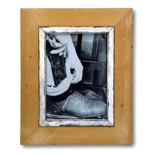 May include: Black and white framed photograph of a person putting on a vintage leather soccer cleat. The frame is a distressed, rectangular shape with a yellow-brown finish and white accents.