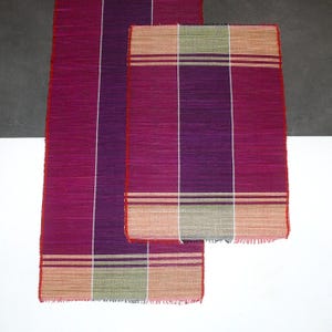 Peut inclure: Deux tapis rectangulaires tissés dans des tons de violet, de rose et de beige. Les tapis présentent un motif rayé avec des accents blancs, beiges et verts. Les bords sont finis avec une bordure rouge et une bordure effilochée. Les tapis sont posés sur une surface blanche et grise.