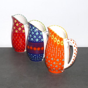 May include: Three ceramic pitchers with colorful geometric patterns. The pitchers are red, blue, and orange with white accents.