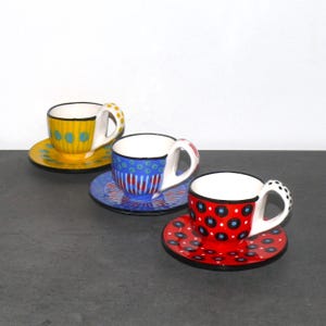 Könnte beinhalten: Drei Espresso-Tassen aus Keramik mit passenden Untertassen und handbemalten Mustern. Eine Tasse ist gelb mit blauen Punkten, eine blau mit roten Streifen und eine rot mit schwarzen und blauen Punkten. Jede Tasse hat ein weißes Innere und einen Henkel.