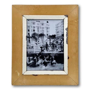 May include: A framed black and white photograph depicting a group of people in a pool, with a coastal city backdrop. The frame is a distressed, light brown color with a white inner border. The photograph captures a vintage scene.