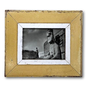 May include: A framed black and white photograph featuring a large stone statue and a person standing on a wall. The frame is rectangular with a distressed yellow exterior and a white interior border.