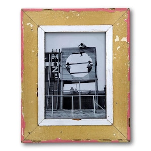 May include: A framed black and white photograph featuring a construction scene. The frame has a distressed look with a gold inner border, a white inner frame, and a pink outer edge. The photograph depicts workers on a wooden structure.