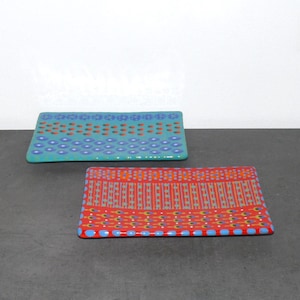 Può includere: Due vassoi rettangolari in ceramica con motivi geometrici colorati. Il vassoio superiore è turchese con accenti rossi, verdi e blu. Il vassoio inferiore è rosso con accenti gialli, verdi e blu.