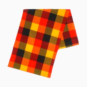 May include: A rectangular, folded fabric with a vibrant plaid pattern. The design features a grid of large squares in shades of red, orange, yellow, and black. The fabric is likely a scarf or a similar textile.
