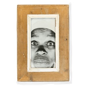 May include: A black and white portrait framed in a rustic wooden frame with a white inner border. The portrait features a person's face with wide eyes and a surprised expression, creating a striking visual contrast.