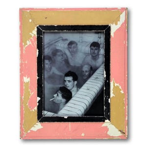 May include: A framed black and white photograph of several men in a pool, framed by a distressed pink, gold, and black wooden frame. The photograph shows the men in the water, some with cigarettes.