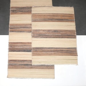 May include: Two woven placemats with a brown and beige striped pattern. The placemats are made of natural materials and have a fringed edge.