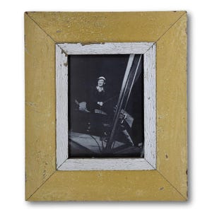 May include: A vintage photograph in a distressed yellow and white wooden frame. The black and white photo features a person in a hat, seated and holding a rope. The frame has a rustic, aged appearance, with visible paint chipping.