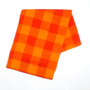 May include: An orange and red checkered fabric, folded and displayed against a white background. The fabric has a bold, vibrant pattern of large squares in shades of orange and red. The fabric is likely a kitchen towel or decorative cloth.