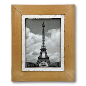 May include: A framed black and white photograph of the Eiffel Tower in Paris. The frame is rectangular with a distressed, aged look, featuring a gold-toned exterior and a white interior border. The image captures the iconic landmark against a cloudy sky.