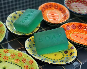 May include: Two rectangular translucent green bars of soap, each embossed with the word SOAP, are resting on individual oval ceramic soap dishes. The dishes feature colorful hand-painted patterns in yellow, orange, and green with small drainage holes.