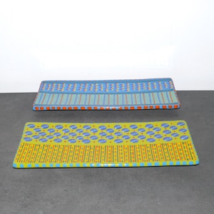 Peut inclure: Deux plateaux de service rectangulaires en céramique avec des motifs géométriques colorés. Le plateau du haut est bleu avec des accents orange, jaune et vert. Le plateau du bas est vert avec des accents bleus, orange et jaunes.