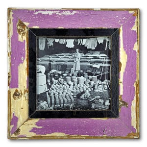 May include: Square framed black and white photograph with a distressed purple and gold frame. The photograph depicts a vintage scene with a woman, stacks of dishes, and hanging laundry.