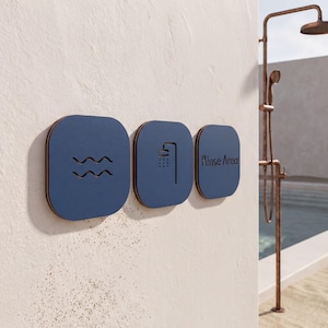 Könnte beinhalten: Drei quadratische blaue Wandschilder mit abgerundeten Ecken an einer strukturierten cremeweißen Wand. Das erste Schild zeigt ein Wellen-Symbol, das zweite ein Duschkopf-Symbol und das dritte den Text Rinse Area in einer dunklen Schrift.