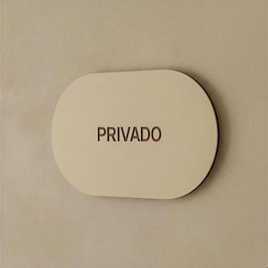 Könnte beinhalten: Ein ovales Schild mit einer glatten, hellbeigen Oberfläche, auf dem das Wort PRIVADO in dunkelbraunen Großbuchstaben eingraviert ist. Das Schild hat ein klares Design mit abgerundeten Kanten und ist flach an einer hellbeigen Wand montiert.